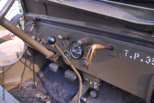 Vintage military vehicle dashboard control panel. Round speedometer and tachometer gauges visible. Wooden steering wheel with metal control levers. 