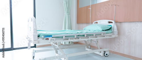 A hospital room shows an empty bed ready for a patient. The bed has light blue sheets and there are some medical devices on the wall. Bright light comes through the window.