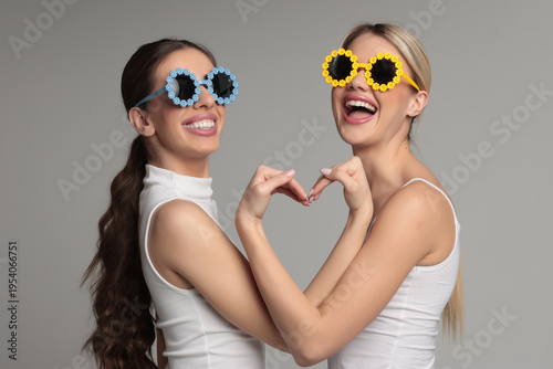 Studio shot of two young women best friends wearing funny party glasses laughing and making heart shape gesture on gray background
