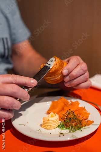 A guest spreads butter on a flaky croissant beside smoked salmon, butter rosette, and microgreens on an orange table in a luxury hotel restaurant in Oberlech, Austria.