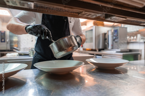A chef in black gloves plates food from a small saucepan onto white dishes in a stainless steel kitchen in Oberlech, Austria, with warm focused lighting and a clean pass counter.