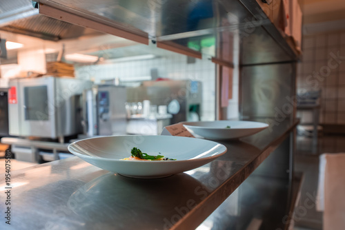 Two plated dishes rest on a stainless pass under heat lamps in a hotel kitchen in Oberlech, Austria. Ovens line the space as soft daylight reflects off metal surfaces.