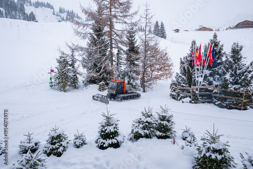 A red and gray snow groomer sits by fir and larch trees, fresh tracks mark a ski slope, flagpoles with national flags stand near chalets in Oberlech, Austria, snowfall