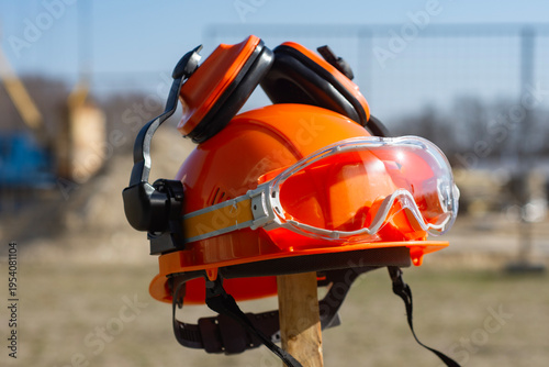 Foto Orange hard hat equipped with safety glasses and noise-canceling ear protectors placed on a wooden stake