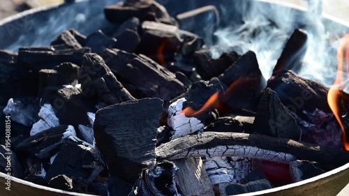 harcoal embers in a cast-iron fire pit, showing the transition from black wood coal to white ash, with glowing orange flames licking through the pile and wisps of white smoke rising in bright daylight