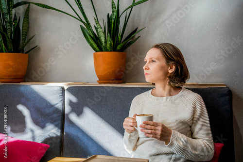 A thoughtful woman in a white knitted sweater sits in a cafe, holding a coffee cup. Peaceful atmosphere in a bright, modern interior.