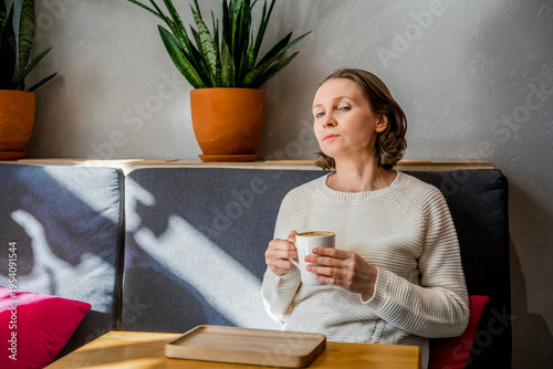 A thoughtful woman in a white knitted sweater sits in a cafe, holding a coffee cup. Peaceful atmosphere in a bright, modern interior.