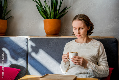 A thoughtful woman in a white knitted sweater sits in a cafe, holding a coffee cup. Peaceful atmosphere in a bright, modern interior.