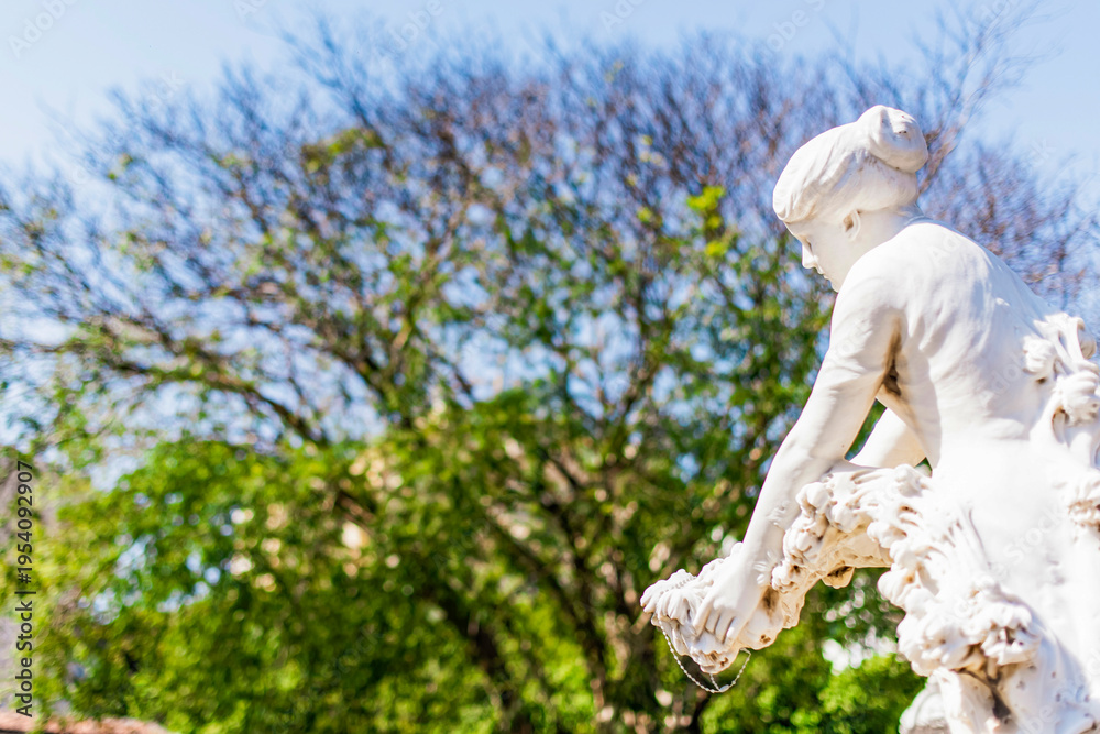 Obraz premium Female sculpture superimposed on a background filled with blurred trees.Sunlight highlights the marble,creating a vibrant contrast with the landscape.Artist unknown, São João Batista Cemetery, Brazil 