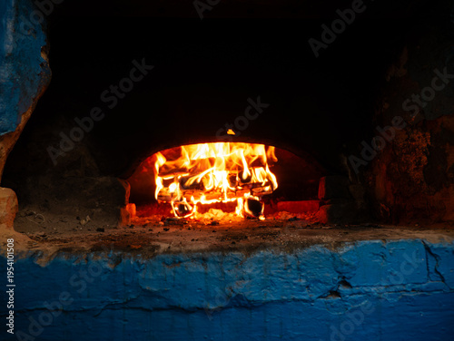 Bright orange flames consume firewood logs inside dark arched chamber of rustic blue painted stone oven during traditional baking process.