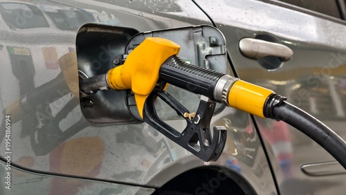 Fotografie A yellow fuel nozzle is inserted into the gas tank of a gray car at a fueling station, highlighting the routine process of refueling a modern internal combustion engine vehicle for transportation