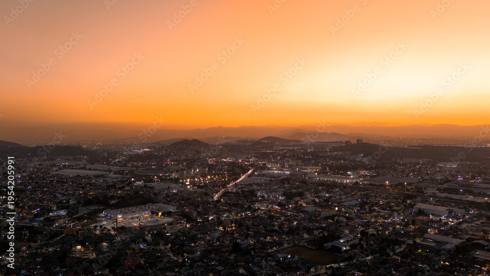 Obraz premium Aerial cityscape at sunset with glowing orange sky and illuminated urban lights
