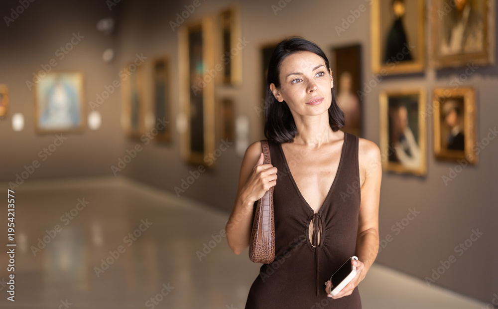 Fototapeta premium Young woman in elegant dress stands in the lobby of museum and looks with interest at the paintings in the gold frames. Tourist with a mobile phone in her hands visits an exhibition of paintings
