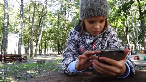 Little girl in a jacket studies the Internet in a mobile mobile phone. City Park. Slow motion.