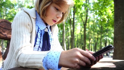 Little girl is sitting in the park at the table and play with a smartphone.
