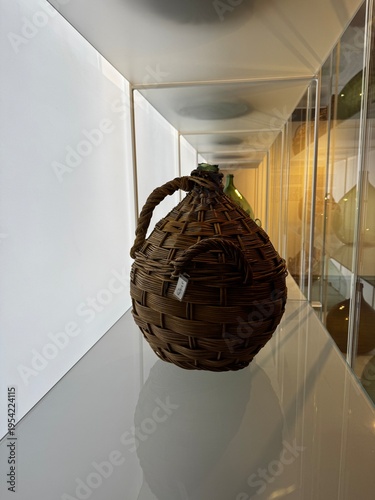 A traditional woven basket container exhibited inside a sleek glass museum showcase. The handcrafted texture and organic material contrast beautifully with the modern minimal display environment, high