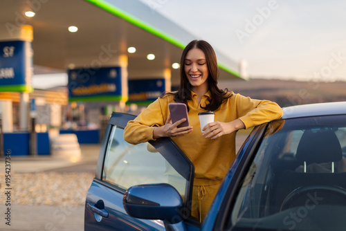 Quadro su tela Lady using cellphone and drinking coffee while stopped at gas station, standing