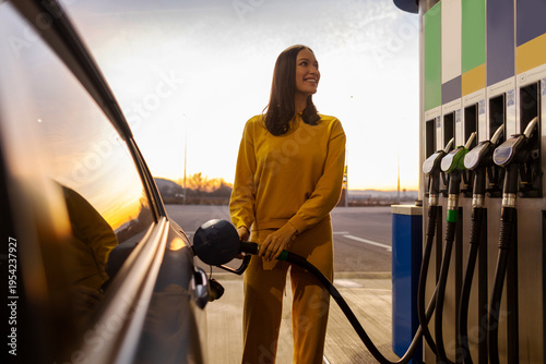 Woman driver refueling her car at petrol station, standing near vehicle holding filling gun nozzle and looking at gasoline dispenser machine
