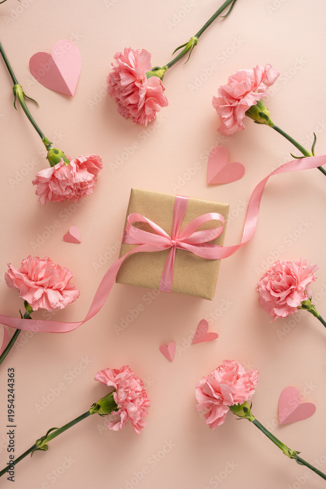 Naklejka premium Happy vertical Mother's Day concept featuring a wrapped gift with pink ribbon, a single carnation flower and paper hearts on a soft peach pastel background ideal for greeting cards
