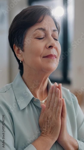 A serene older woman meditates with eyes closed and hands clasped in prayer, in a peaceful indoor setting with a blurred background