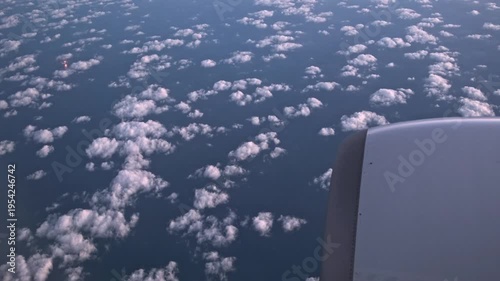 airplane window captures small cumulus clouds over the dark ocean at dusk and A bright orange light source, likely a ship, oil platform