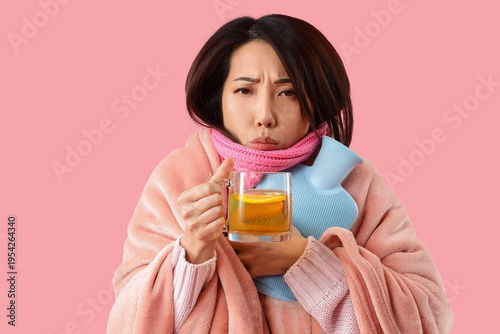 Canvas Print Ill young Asian woman with hot water bottle and glass cup of green tea on pink b