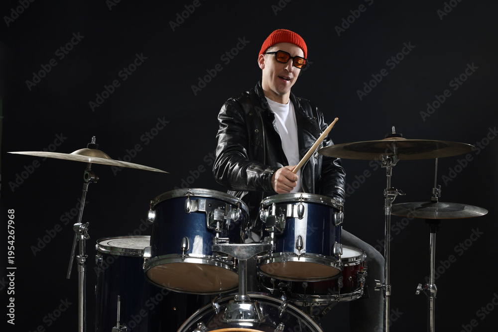 Obraz premium Smiling man in hat and sunglasses playing drums with drumsticks on black background