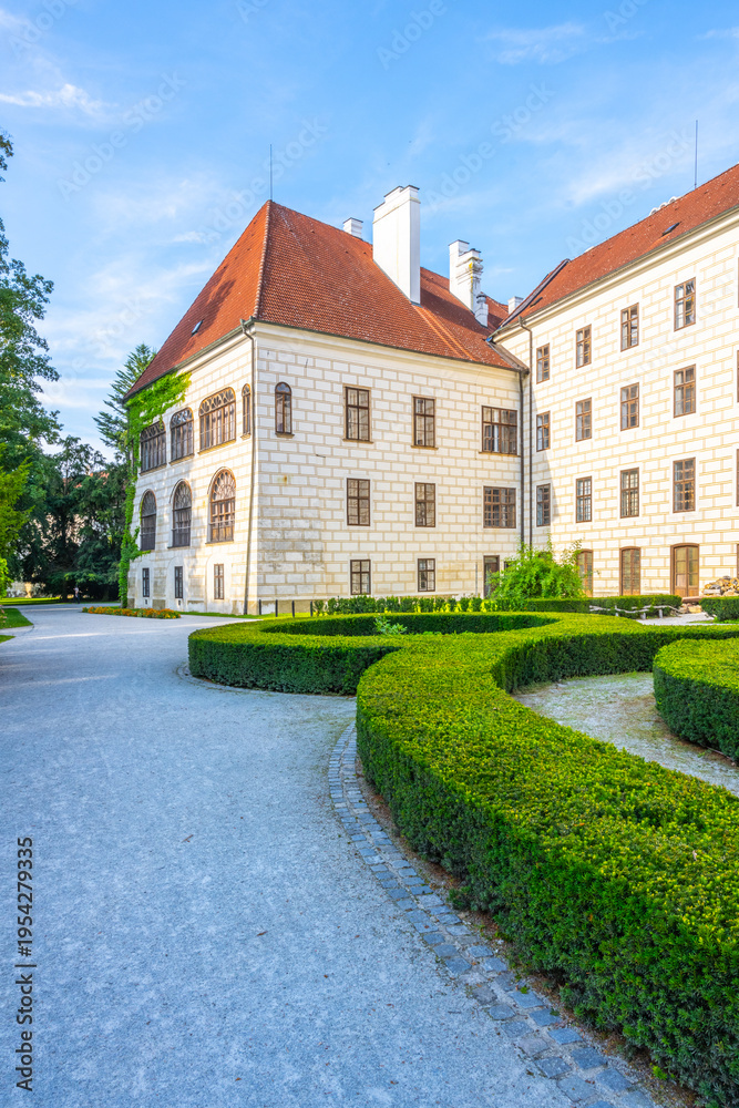 Naklejka premium Trebon Castle stands in the background while visitors walk along the path. The garden is well kept with trimmed bushes. Sunlight shines on the stone walls.