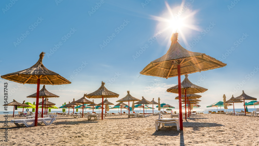 Fototapeta premium Sandy beach with rows of straw umbrellas on the Black Sea resort of Mamaia, Romania, in the morning after the sunrise, with the sun shining above the Black Sea.