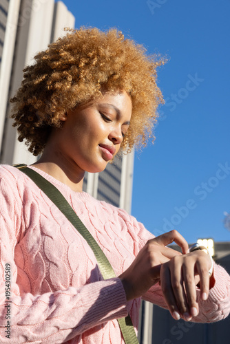 African American woman checking watch on wrist in urban area, wearing pink sweater and green strap