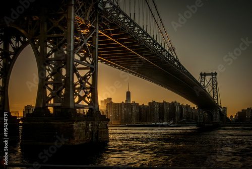 A stunning sunset view under the massive steel structure of the Williamsburg Bridge in NYC, with the Manhattan skyline and One World Trade Center visible in the hazy distance. 