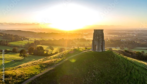 Wallpaper Mural Aerial view captures a sunlit ancient stone tower atop a grassy hill as the sun peeks over a landscape Torontodigital.ca