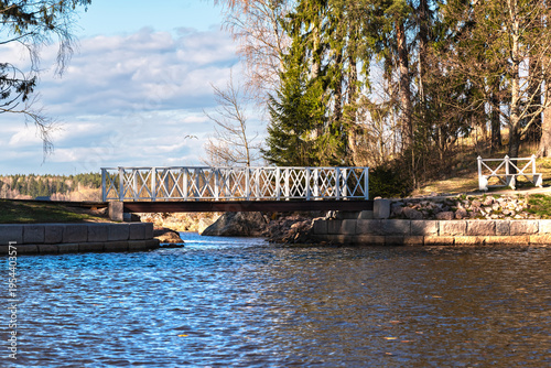Wooden bridge with white railings connecting the two banks across a small channel in the city park on a sunny spring day.
