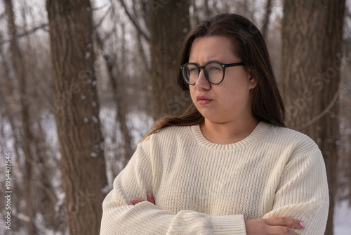 A thoughtful young woman with glasses and a cream sweater stands outside in a snowy forest. With arms crossed, she looks away with a serious, pensive expression. Soft light, muted natural colors.