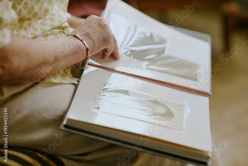 Senior Caucasian woman sitting and browsing photo album, gently turning page with left hand, focusing on black and photographs, capturing nostalgic family memories