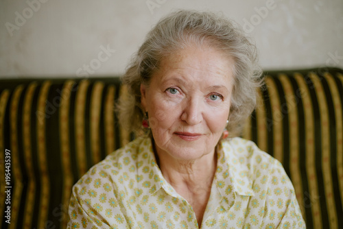 Portrait of senior Caucasian woman sitting on striped sofa looking directly at camera with gentle expression, short gray hair framing face, wearing patterned blouse and earrings