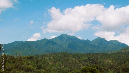 Bilde på lerret A serene mountain range rises above a lush forest under a blue sky with white cl