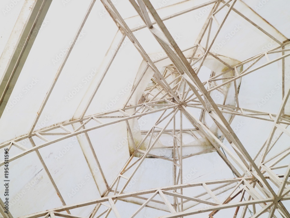 Obraz premium Intricate network of beige metal beams and supports under a transparent roof with a clear blue sky visible