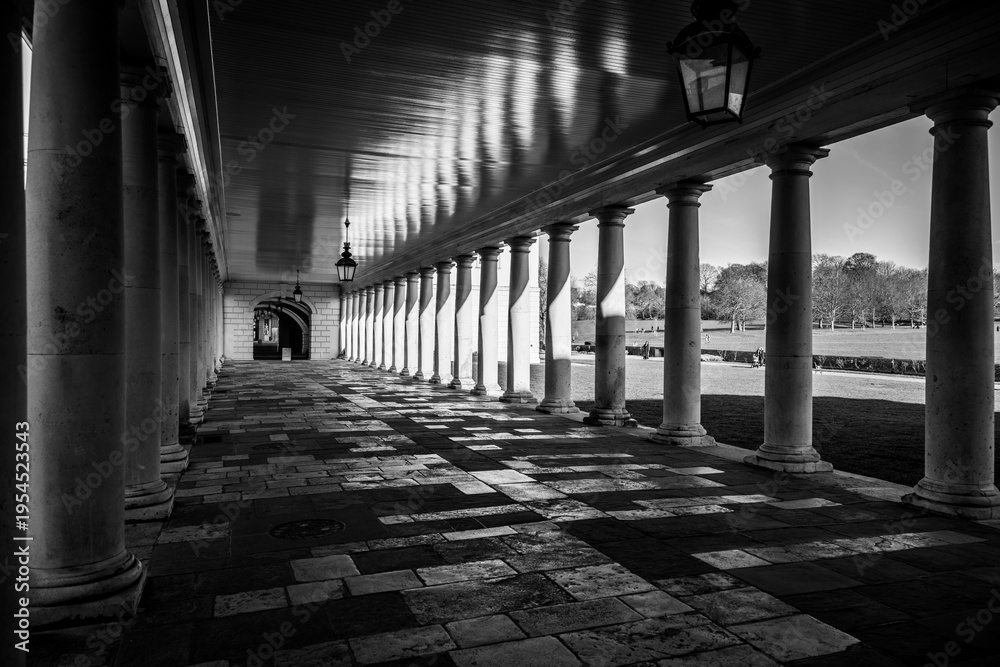 Fototapeta premium Visitors walk through the corridor of Greenwich Queens House in London. The building features classic architecture and a well-maintained path. Sunlight creates shadows on the ground.