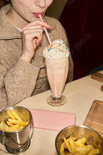 Vertical midsection shot of young woman drinking milkshake with whipped cream and sprinkles through straw while sitting at table with French fries in foreground