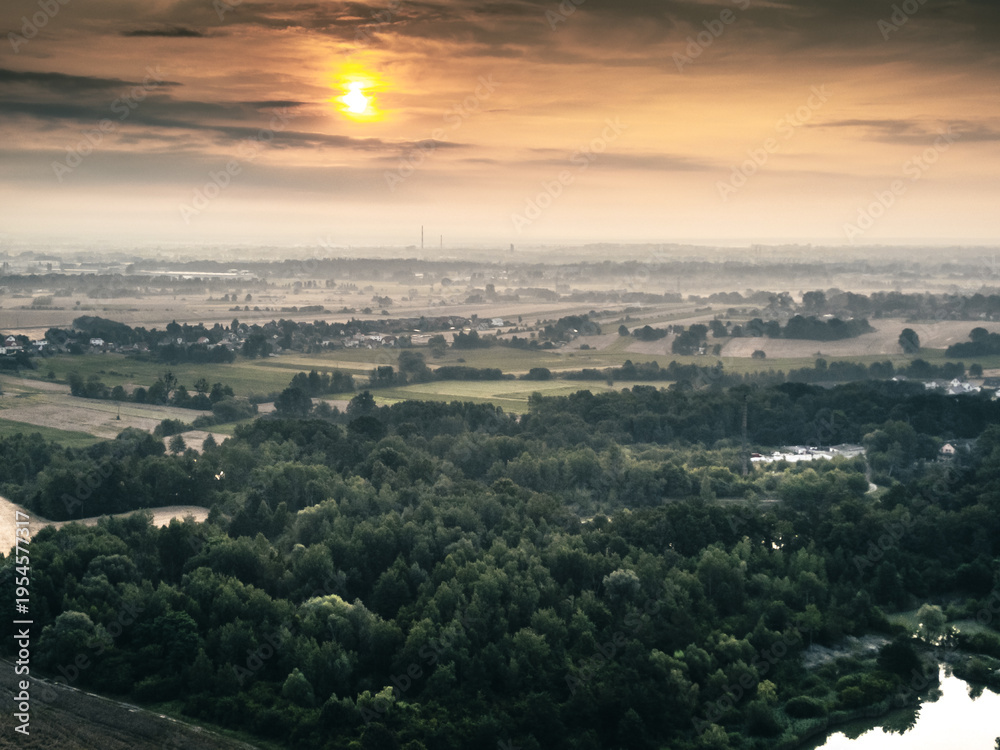 Fototapeta premium Malowniczy zachód słońca nad polskimi polami i lasami. Widok z góry na krajobraz rolniczy, wieś w oddali i nastrojową mgiełkę. Fotografia z drona.