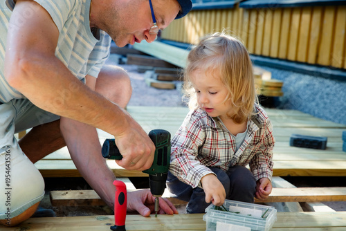 Joyful father and daughter building a wooden patio deck together. Authentic family DIY home improvement project, sunny outdoor leisure.