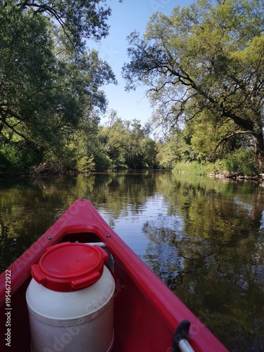 Rotes Kanu auf einem ruhigen Fluss mit grünen Bäumen an den Ufern, Perspektive vom Boot aus (POV)