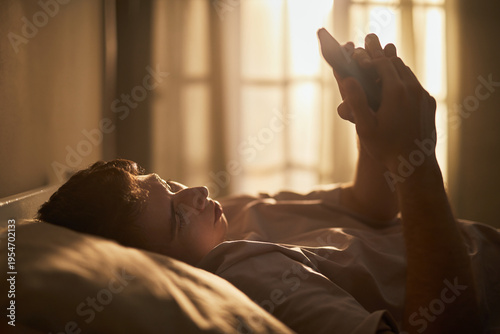 Photography Caucasian teenage boy lying on bed holding smartphone with both hands, gazing at