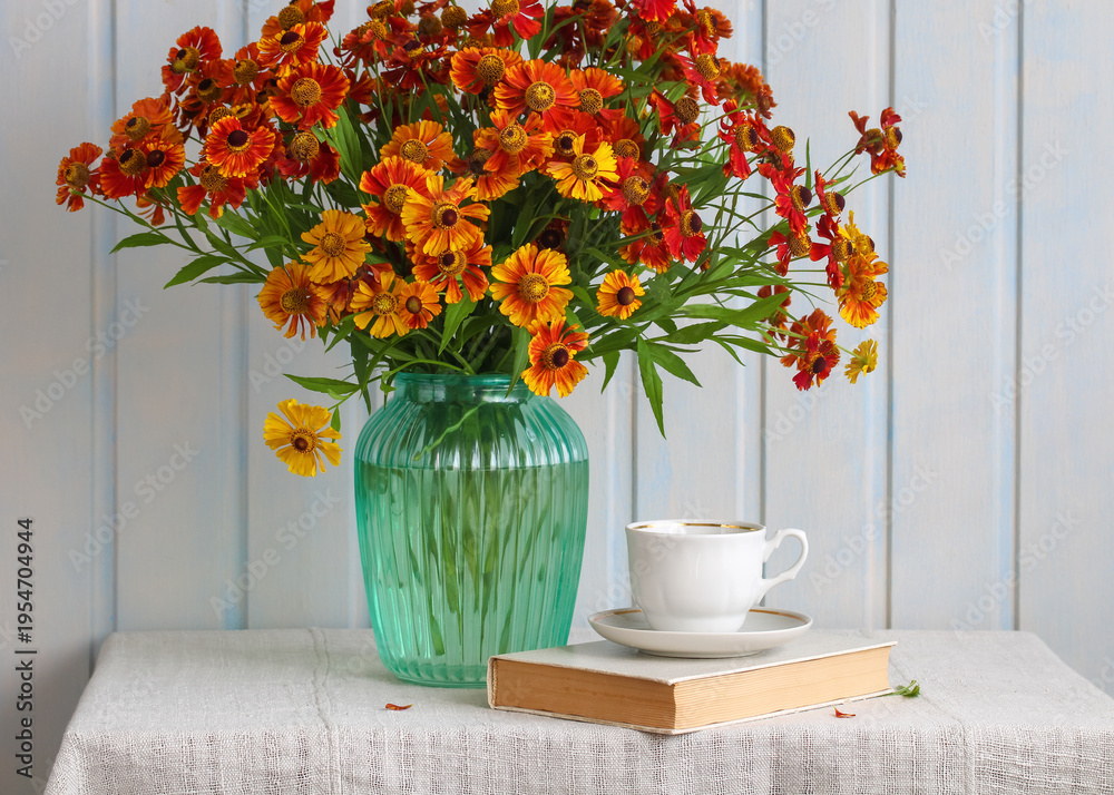 Naklejka premium Vibrant Helenium flowers bouquet filling a teal ribbed glass vase, contrasting with a white porcelain teacup on an antique book, creating a tranquil still life composition