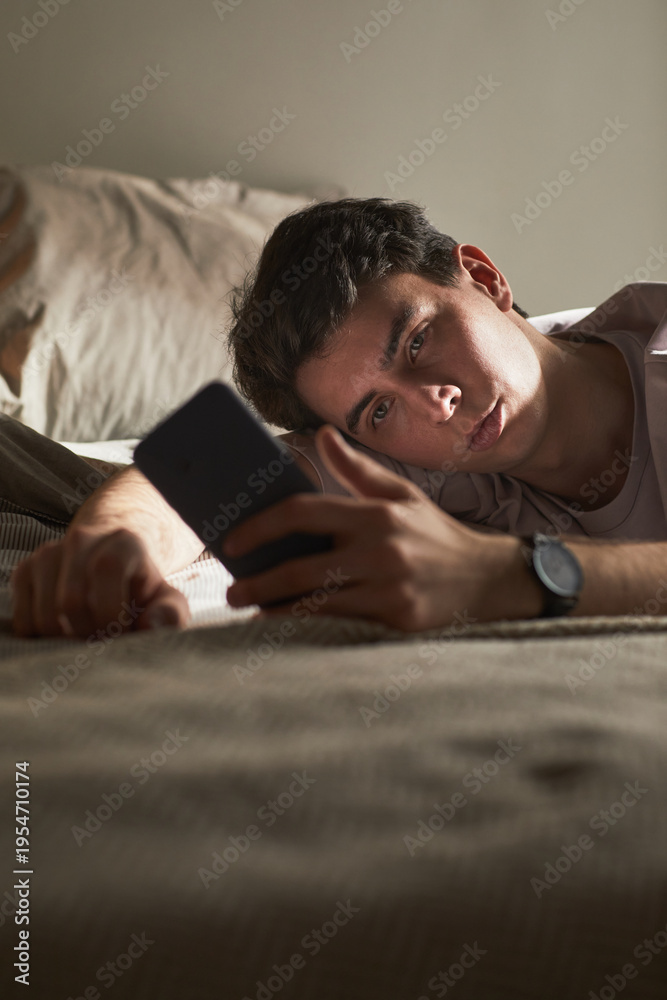 Fototapeta premium Vertical of Young Caucasian man lying on bed holding smartphone, gazing at screen with neutral expression, showing signs of emotional fatigue or contemplation in relaxed indoor setting