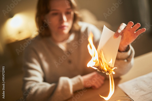 Young adult Caucasian woman burning boyfriend photo with focused expression, sitting at table indoors, holding flaming sheet in hand, demonstrating intense emotion and purposeful action