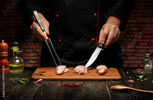 A chef prepares for cooking by cutting chicken pieces on a wooden board. Tools like a knife and tongs are visible. Spices and oil are set around the board in a kitchen