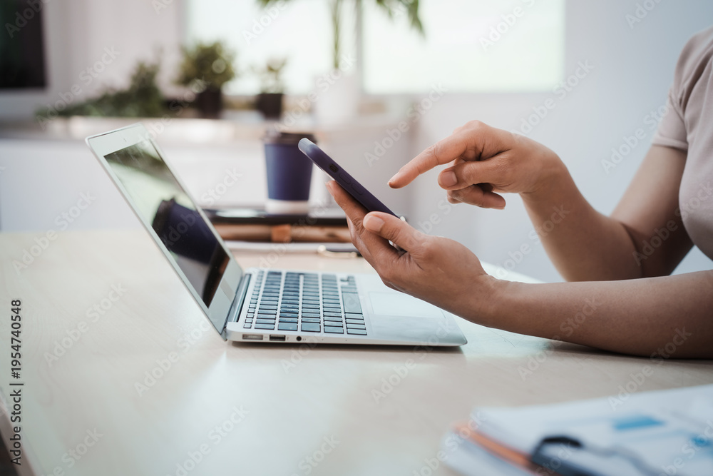 Obraz premium Close-up of person using smartphone beside open laptop on desk in bright modern office, remote work and digital communication concept.