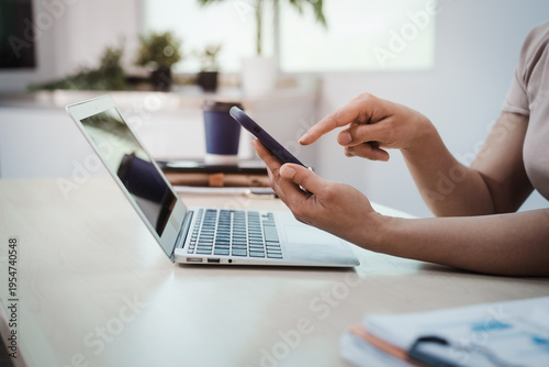 Close-up of person using smartphone beside open laptop on desk in bright modern office, remote work and digital communication concept.